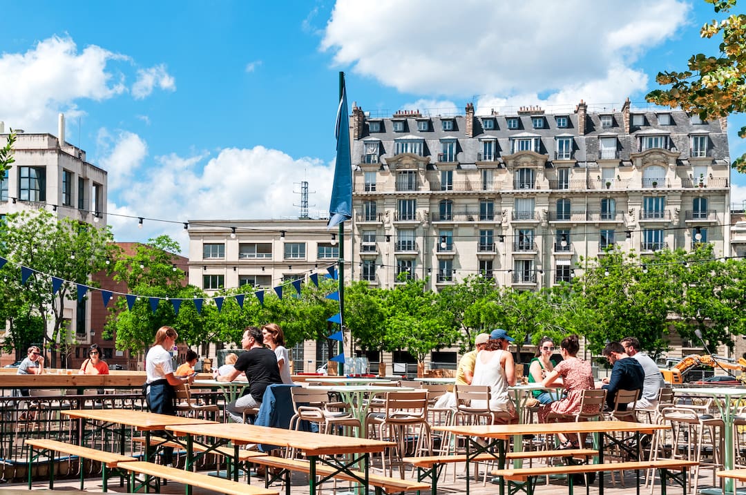 Udendørs café i Bastille-området i Paris, hvor folk nyder en solrig dag ved borde og bænke. Baggrunden viser klassiske parisiske bygninger under en klar blå himmel.