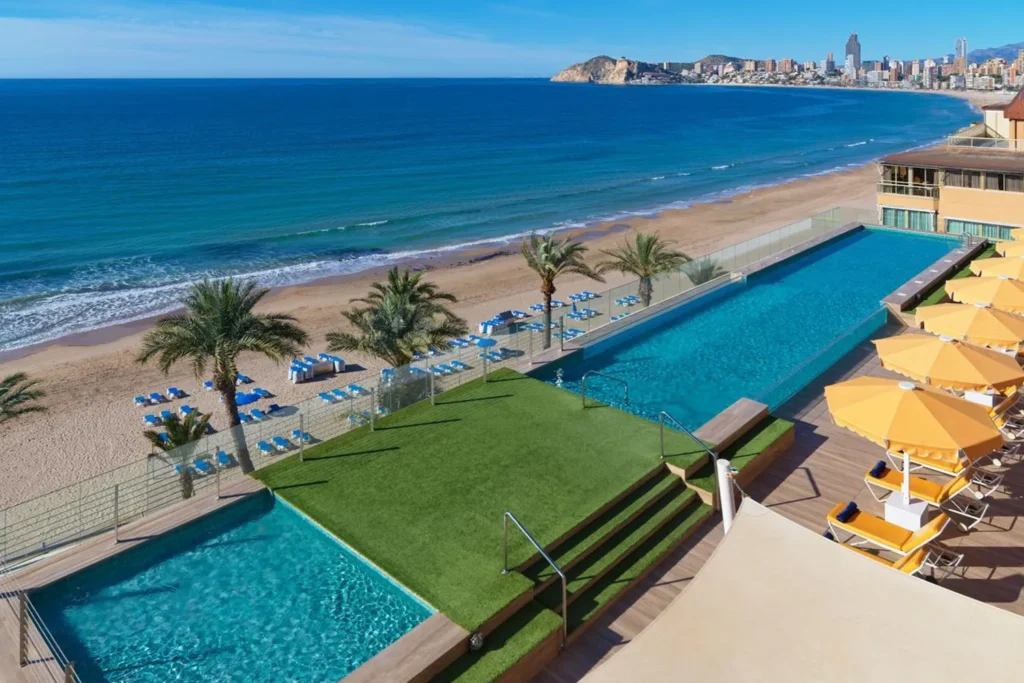 Udsigt over stranden og havet fra Hotel H10 Porto Ponientes rooftop poolområde, med palmer og solstole langs poolen.