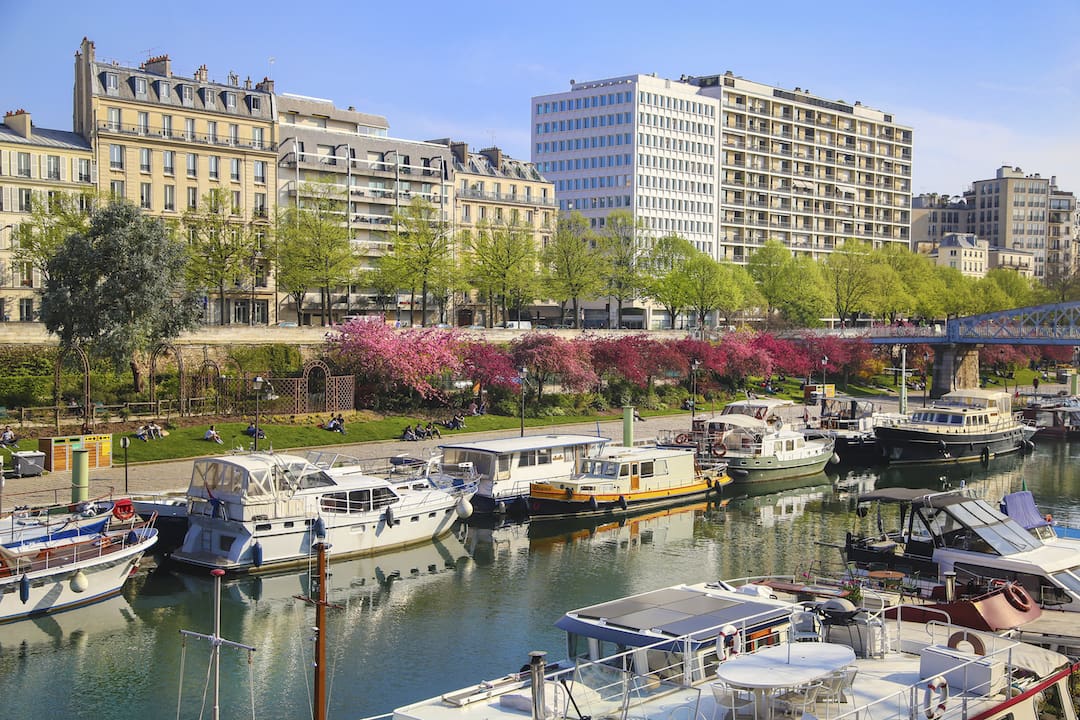Forårsscene langs Le Canal Saint-Martin i Paris, hvor både ligger fortøjet ved kajen, og træerne blomstrer med lyserøde blomster. Folk nyder solen på græsplænerne langs kanalen, omgivet af smukke bygninger og grønne træer
