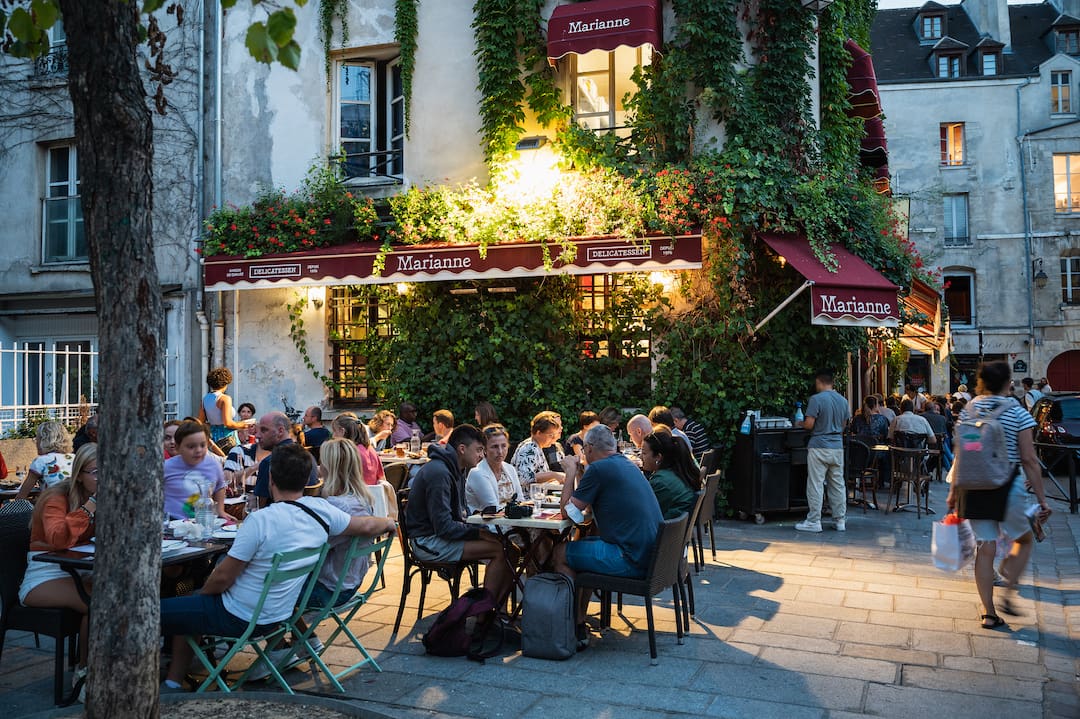 En livlig gadescene i Le Marais, Paris, hvor folk nyder aftenen på en charmerende café med udendørs servering. Bygningen er dækket af frodig grøn beplantning, og caféen har et hyggeligt og afslappet miljø