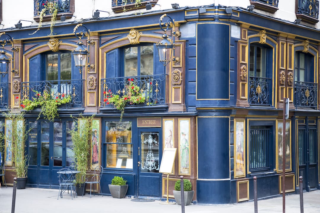 Elegant facade af en bygning i kvarteret Saint-Germain-des-prés, Paris. Bygningen er dekoreret i mørkeblå og gyldne farver med detaljerede ornamenter og blomsterdekorationer på altaner.<br />
