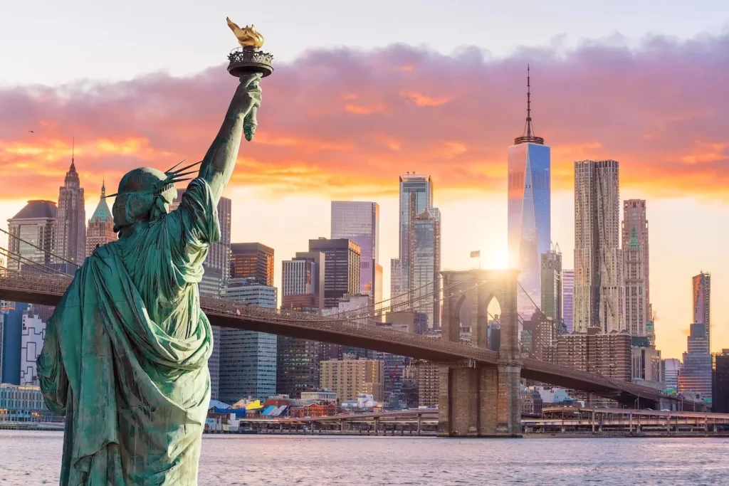 Frihedsgudinden i forgrunden med New York Citys ikoniske skyline og Brooklyn Bridge i baggrunden, oplyst af en smuk solnedgang. Dette billede fanger essensen af New Yorks storhed og tiltrækker rejsende, der søger billige fly til New York for at opleve byens verdensberømte seværdigheder.