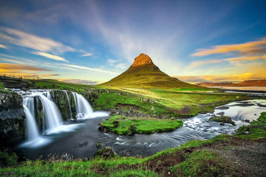 Et fantastisk landskab på Island med det ikoniske Kirkjufell-bjerg i baggrunden og en smuk vandfaldsstrøm i forgrunden, badet i det bløde lys fra den lave sol. Dette maleriske sted fremhæver Islands naturlige skønhed, hvilket gør det til en drømmedestination for rejsende, der søger billige fly til Island.