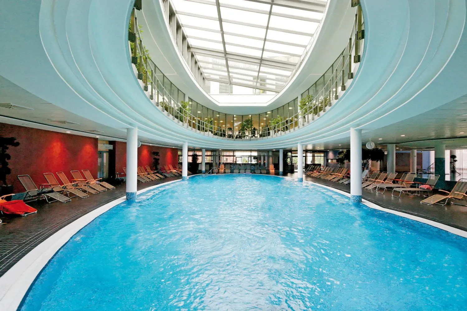 Det indendørs poolområde på Centrovital Hotel Berlin er omgivet af moderne arkitektur med buede balkoner og store glasvægge, der giver masser af naturligt lys. Den runde pool er omgivet af behagelige liggestole, der indbyder til afslapning efter en svømmetur. Det elegante design og den rummelige atmosfære skaber en afslappende wellnessoplevelse i hjertet af Berlin.