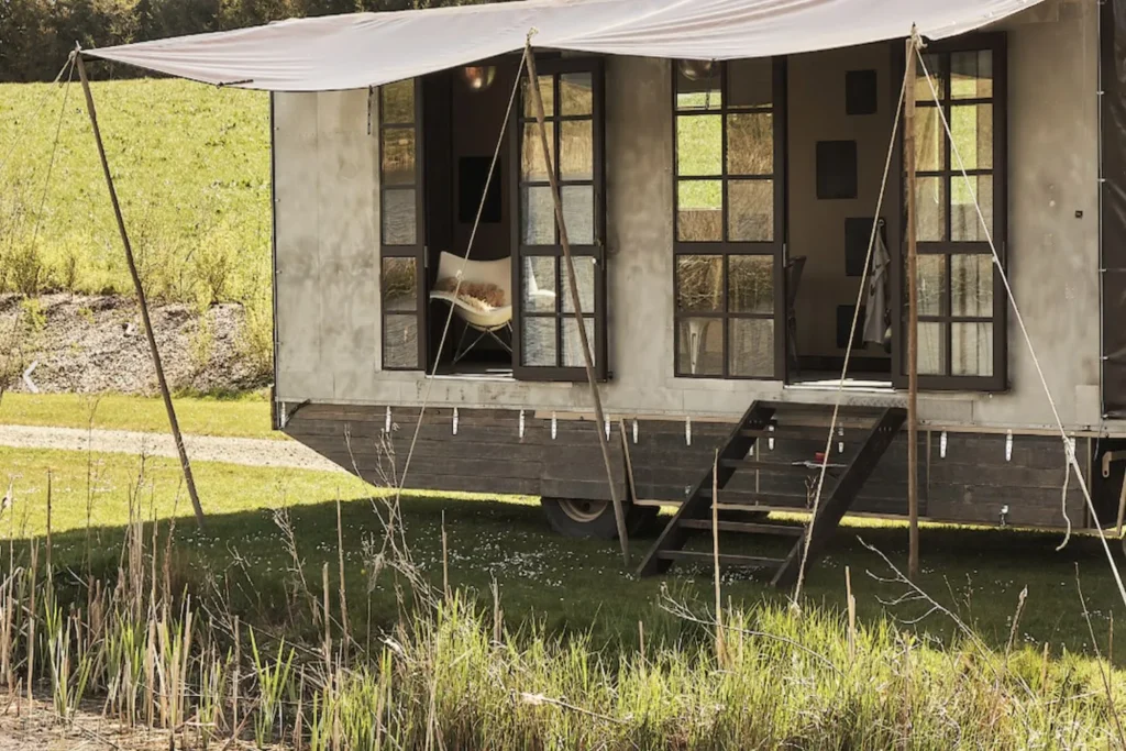 En charmerende cirkusvogn hos Krusmølle Glamping med store vinduer og en overdækket terrasse, der inviterer til afslapning midt i naturen. Vognen står idyllisk placeret ved en lille sø omgivet af grønt landskab, hvilket giver en perfekt kombination af komfort og rustik charme til glampingoplevelsen.