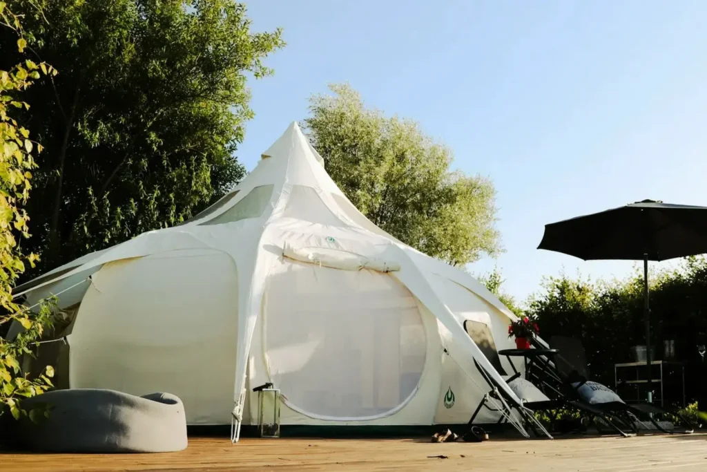 Et rummeligt, hvidt glamping-telt på Thorupgaard, placeret på en træterrasse omgivet af grønne træer og buske. Teltet er stilfuldt og moderne med liggestole og en parasol, der inviterer til afslapning under åben himmel. Den naturskønne placering giver en fredelig og afslappende glampingoplevelse i det danske landskab.