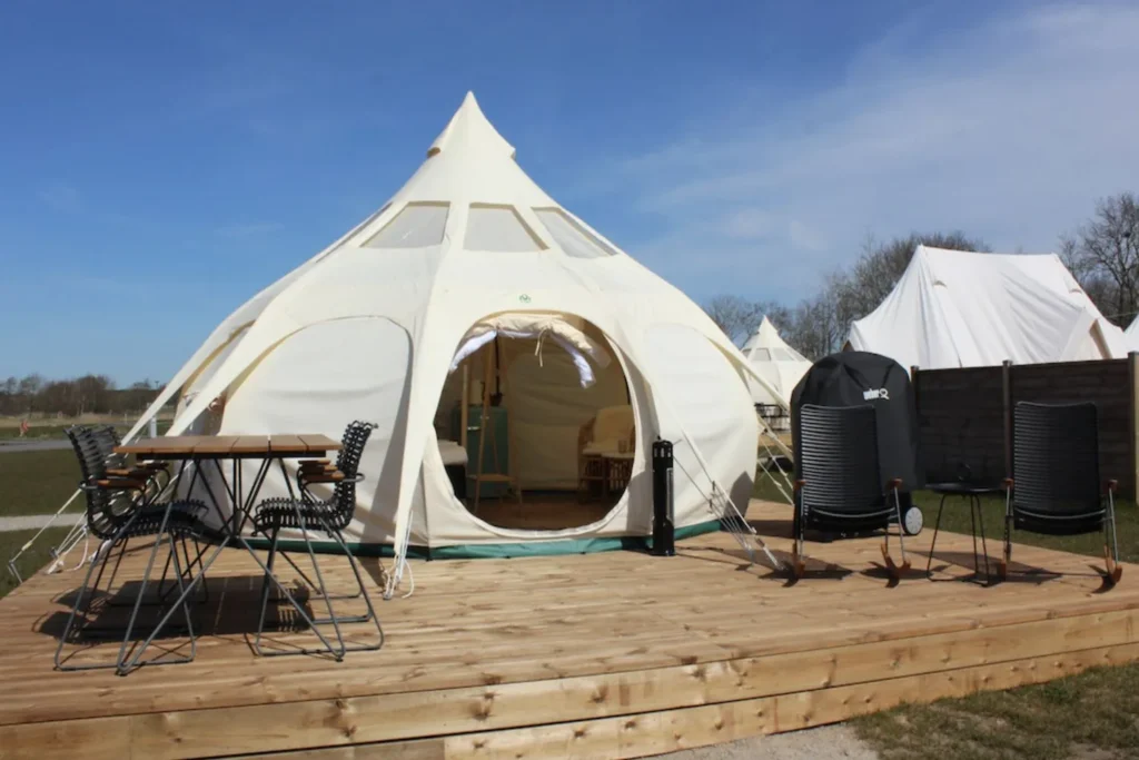 Et rummeligt, hvidt telt på en træterrasse tilbyder en komfortabel glampingoplevelse hos Marsk Camp. Teltet er stilfuldt indrettet og har en hyggelig siddeplads udenfor med bord og stole, perfekt til at nyde udsigten over det åbne landskab. Glamping-teltets unikke form og udendørs faciliteter giver en afslappende og luksuriøs campingoplevelse i naturskønne omgivelser.