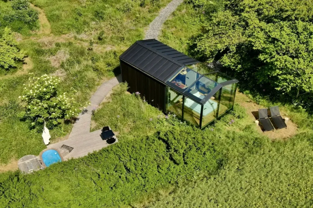 Luftfoto af Glashytterne, hvor en hytte med glasvægge ligger midt i det grønne landskab omgivet af natur. Der er også en udendørs jacuzzi ved siden af hytten.