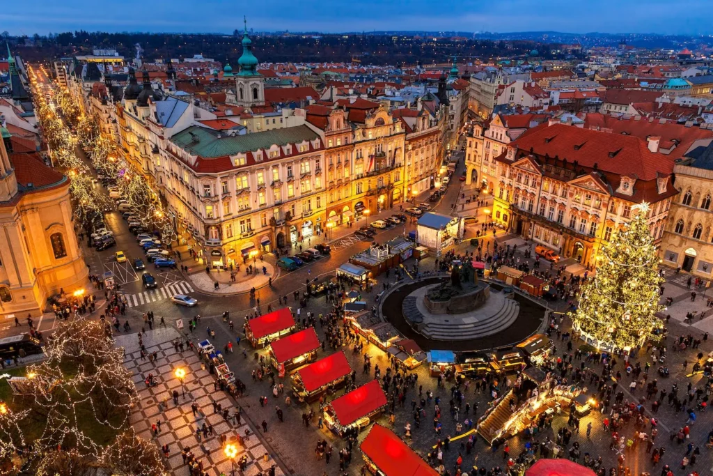 Billede af en livlig julemarkedsscene i hjertet af Prag, hvor smukke, historiske bygninger omkranser det oplyste torv. Røde boder med julepynt og lysende træer skaber en magisk atmosfære, der gør en juletur til Prag til en uforglemmelig oplevelse."