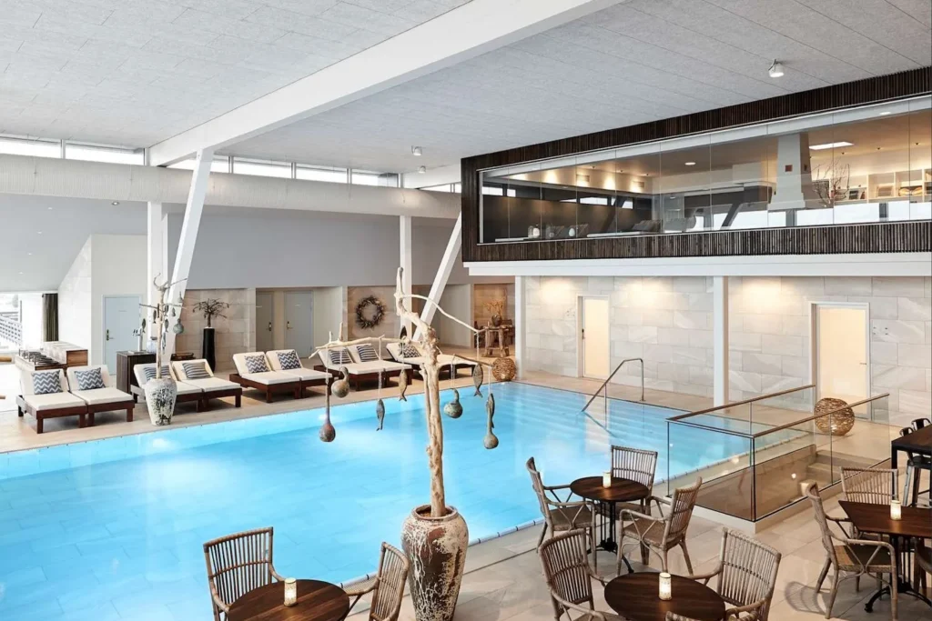 Det elegante spaområde på Marienlyst Strandhotel med en opvarmet indendørs pool omgivet af komfortable liggestole og moderne indretning. Omkring poolen er der hyggelige siddeområder med træborde og kurvestole, hvor gæsterne kan nyde en afslappende atmosfære. Det store vinduesparti på første sal giver masser af naturligt lys, hvilket skaber en lys og indbydende stemning i dette luksuriøse wellnessområde.