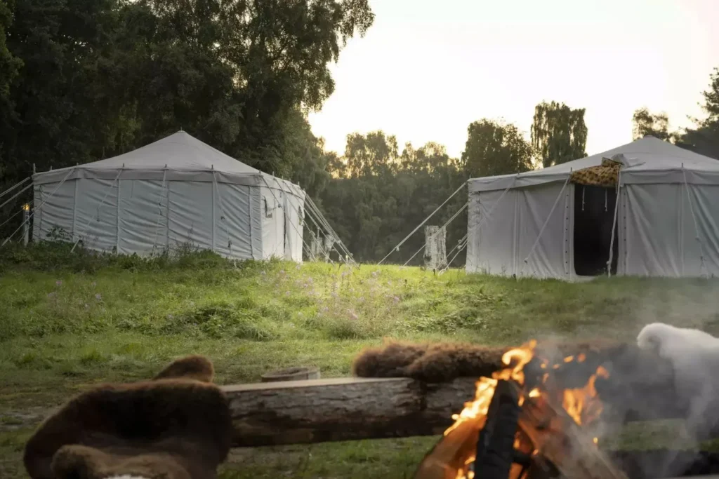 To store, marokkanske glampingtelte står på en åben mark omgivet af skov. Foran teltene er et bålsted med brændende flammer, som skaber en hyggelig og autentisk campingatmosfære. Teltene er rummelige og holdt i en traditionel marokkansk stil, der giver en eksotisk glampingoplevelse midt i naturens ro og skønhed.