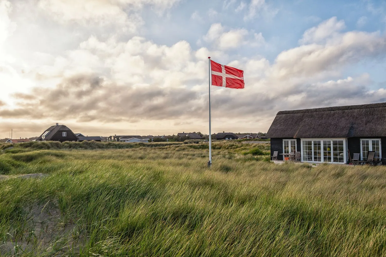 Charmerende sommerhus i klitterne med Dannebrog vajende i vinden under en let overskyet himmel. Den idylliske scene er perfekt til en afslappende miniferie i Danmark, hvor ro og naturskønne omgivelser skaber den ideelle ramme for et pusterum fra hverdagen.