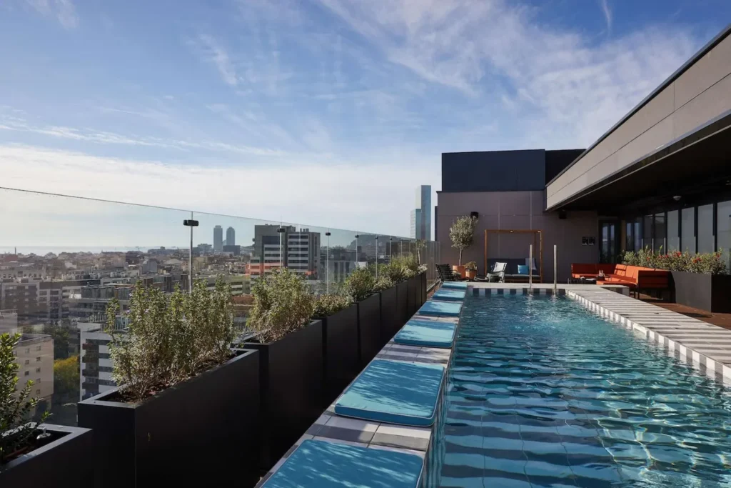 Udsigten fra en tagterrasse under en rejse til Barcelona, hvor en moderne infinity-pool strækker sig langs kanten, omgivet af liggestole og frodige planter. Poolen giver en fantastisk panoramaudsigt over Barcelonas skyline, med ikoniske bygninger, der rager op mod himlen i baggrunden. Dette luksuriøse rooftop-område kombinerer afslapning og byliv, perfekt til at nyde solen og byens livlige atmosfære.