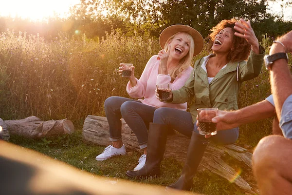 To kvinder griner og nyder drinks i solnedgangen under en hyggelig glamping-oplevelse i naturen.